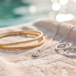 Les bijoux Jolie Demoiselle résistent-ils vraiment à l’eau ? Gros plan de bijoux dorés et argentés disposés sur une serviette de plage, avec une eau claire en arrière-plan et une lumière naturelle douce.